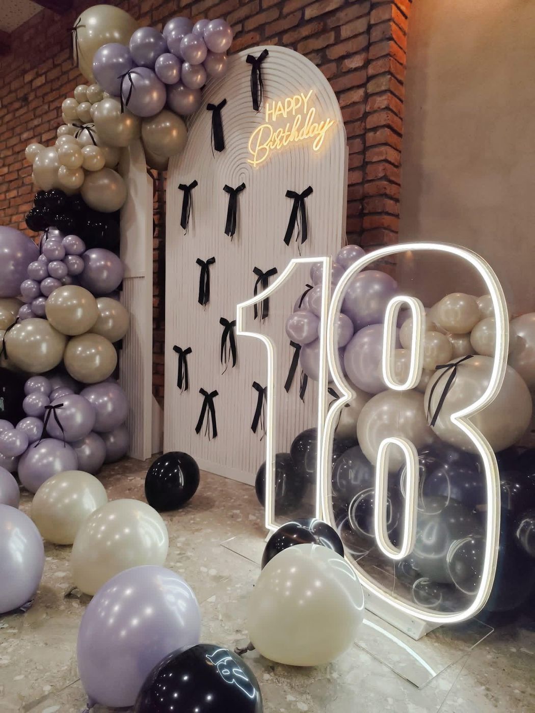 Cifra 18 luminoasă lângă un panou cu fundiţe negre şi baloane lila, sub un semn „Happy Birthday”