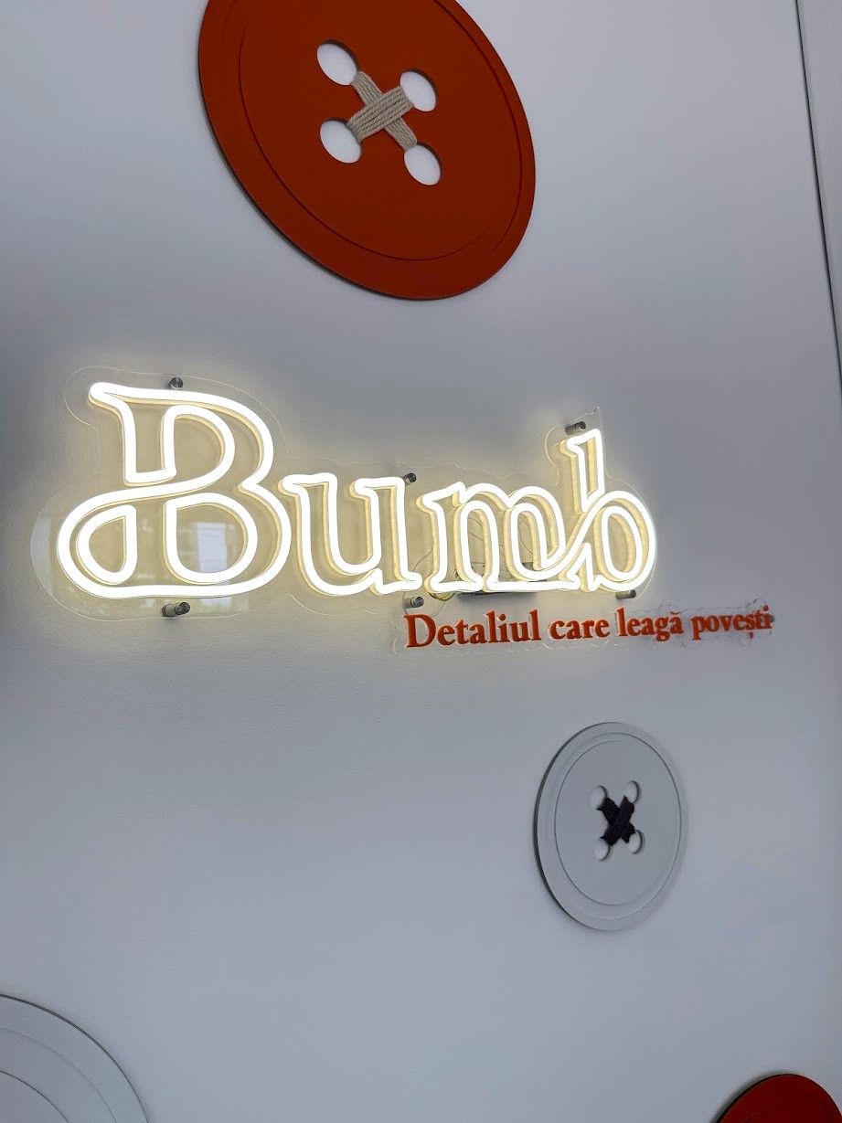 Firmă luminoasă „Bumb — Detaliul care leagă poveşti”, pe un perete alb cu nasturi decorativi