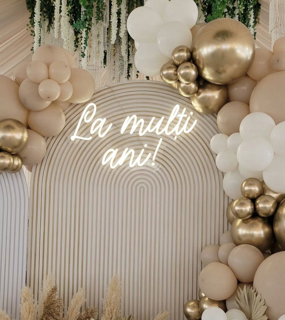 Semn luminos „La mulţi ani!” pe o arcadă crem, între baloane aurii şi pampas